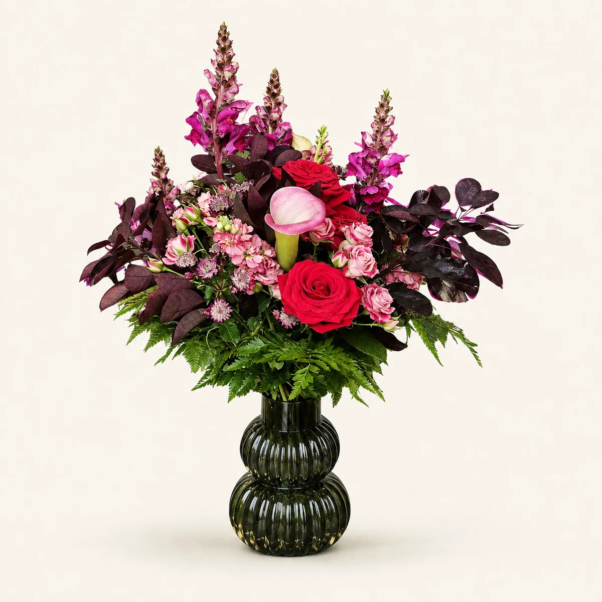 Red roses, a pink calla lily and purple snapdragons with burgundy foliage in a dark green ribbed vase