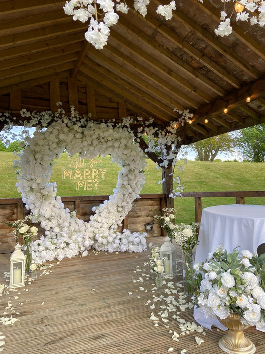 White floral proposal heart with a neon sign, lanterns, candles and floral arrangements in urns.
