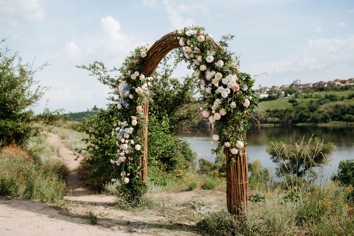 Rustic curved wedding arch decorated with white flowers and trailing greenery beside a lake.