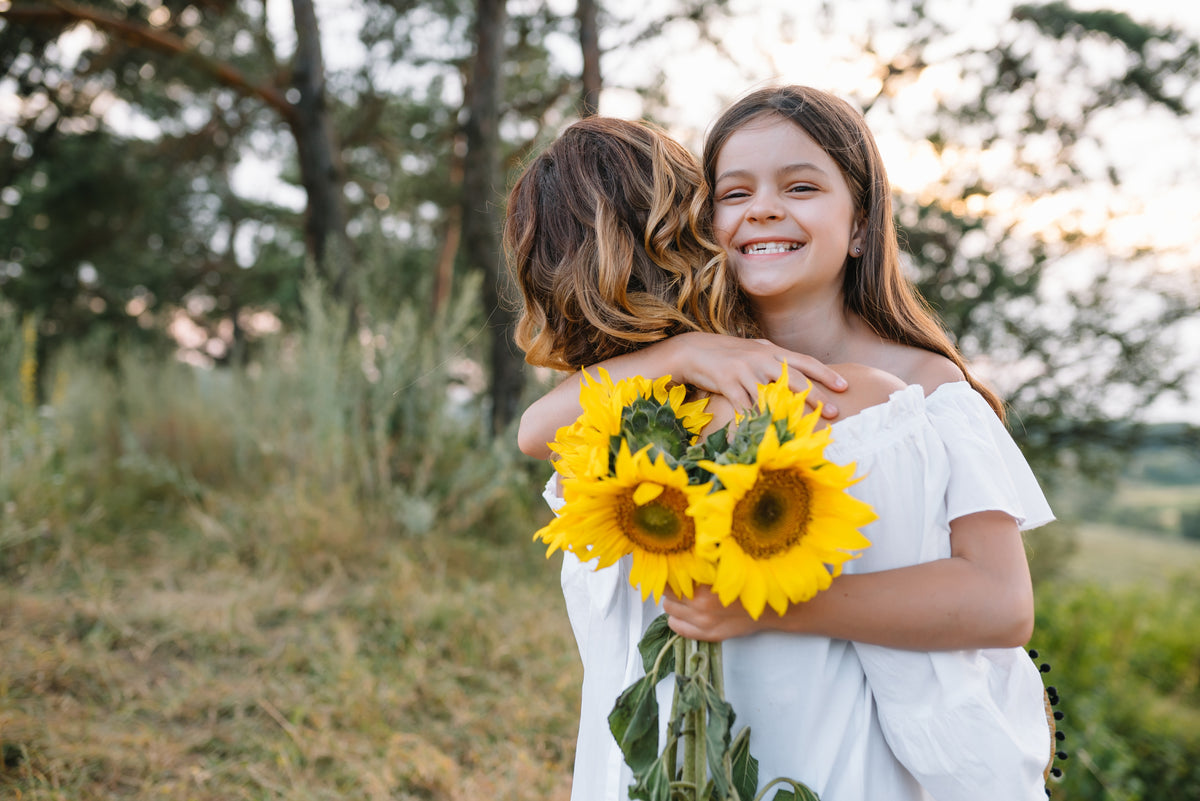 Smiling girl hugging a woman holding a bright yellow sunflower bouquet outdoors.