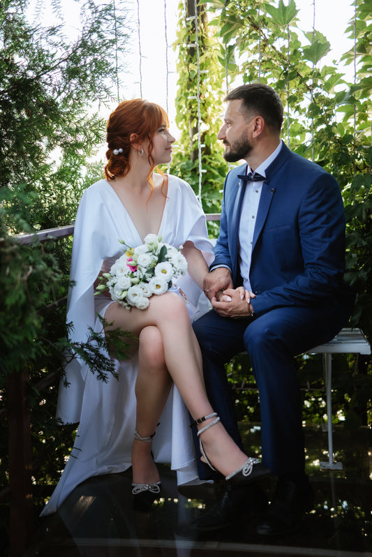 Bride holding a white flower bouquet beside a groom in a blue suit, surrounded by greenery.