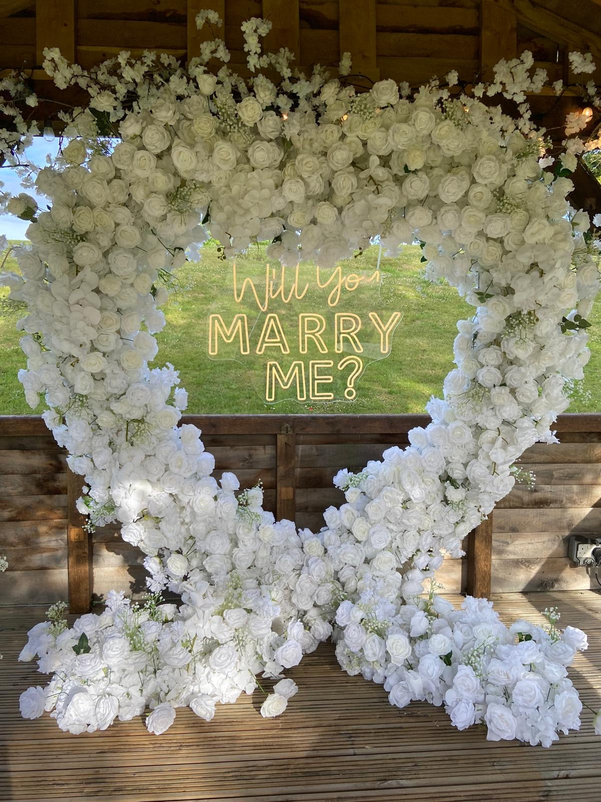 White silk floral heart with an illuminated Will you marry me sign in a wooden gazebo.