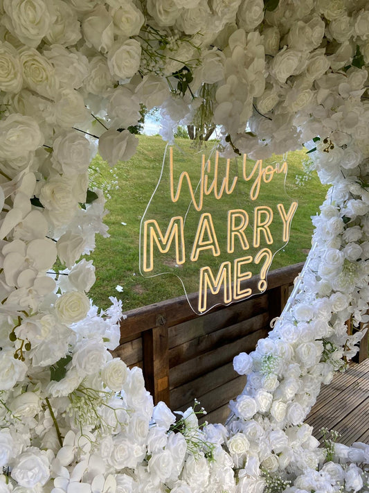 Close-up of a white floral heart surrounding an illuminated Will you marry me sign.
