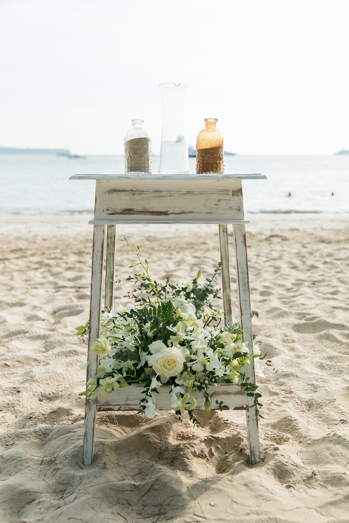 White roses and greenery decorating a wooden stand at a beach wedding ceremony.