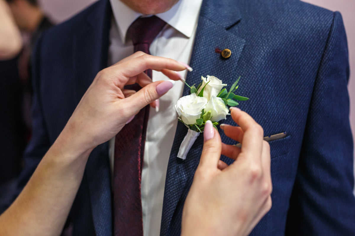 White rose buttonhole being pinned to the lapel of a blue wedding suit.