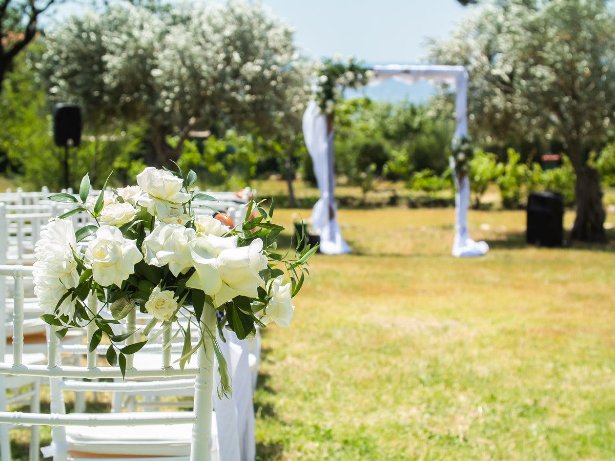 White rose and foliage arrangements on ceremony chairs beside an outdoor wedding aisle.