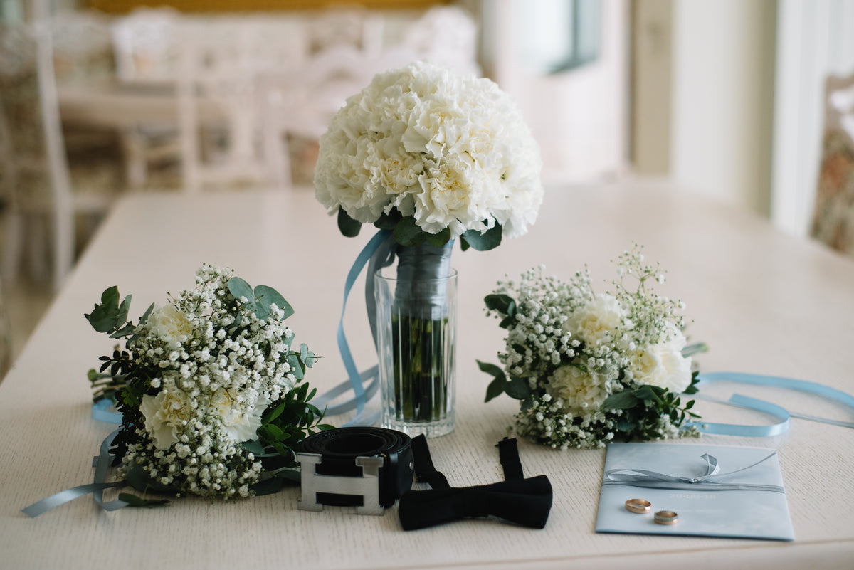 White bridal and bridesmaid bouquets with pale blue ribbons, beside wedding rings and stationery.