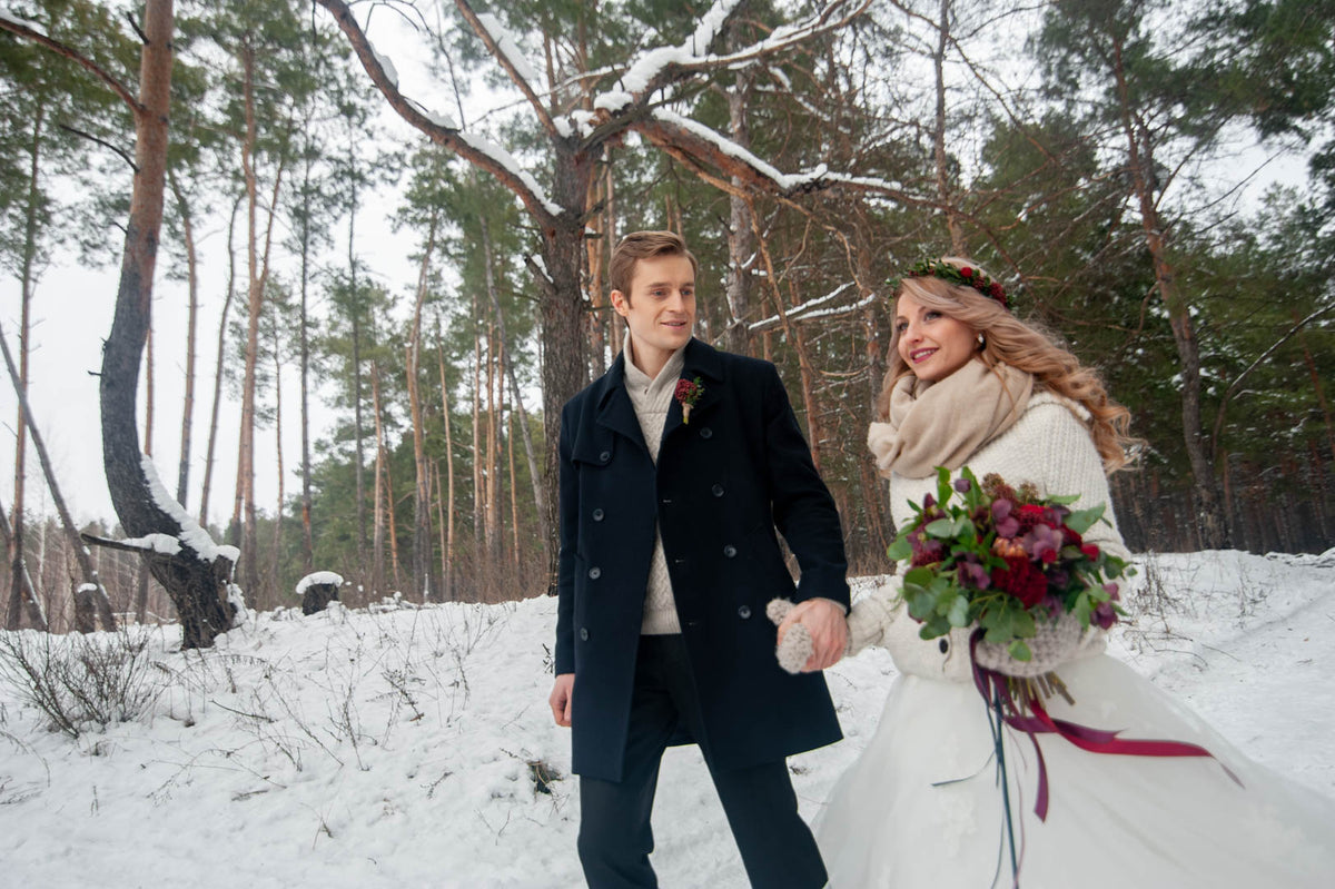 Bride and groom walking in a snowy woodland, with a rich burgundy and green bridal bouquet.