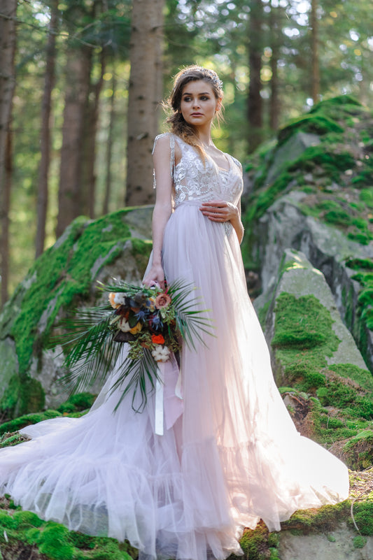 Bride in a pale dress holding a loose bouquet with trailing greenery in a woodland setting.